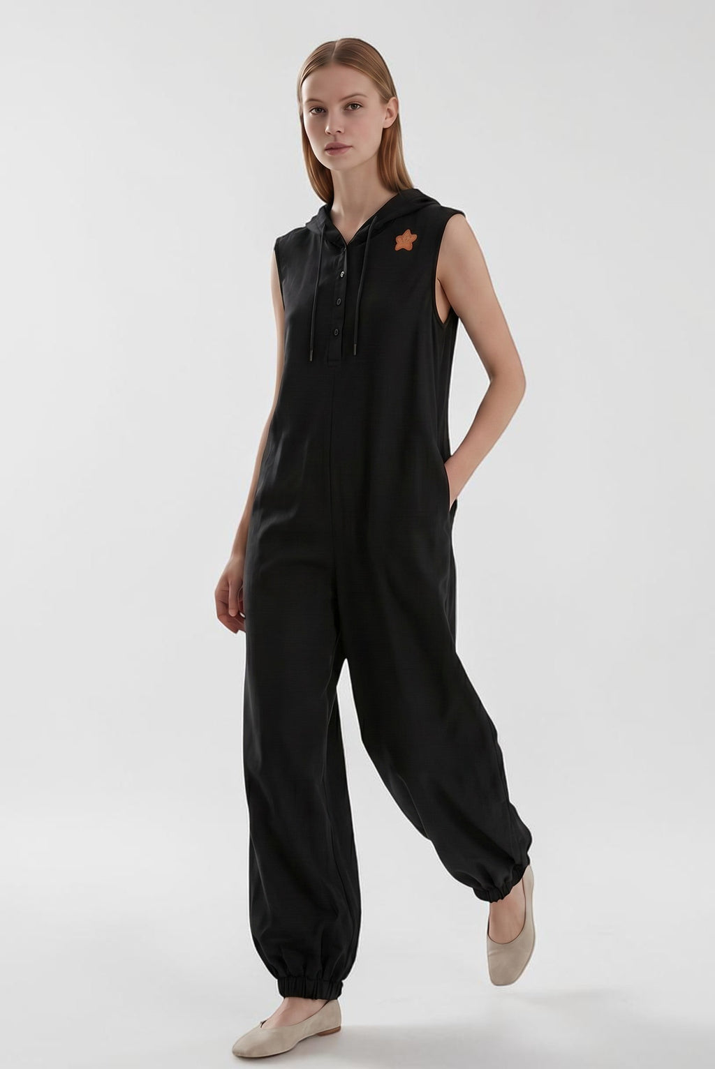 Day One Concept Store black sleeveless wide-leg jumpsuit with hood and star detail. Model wears beige flats.