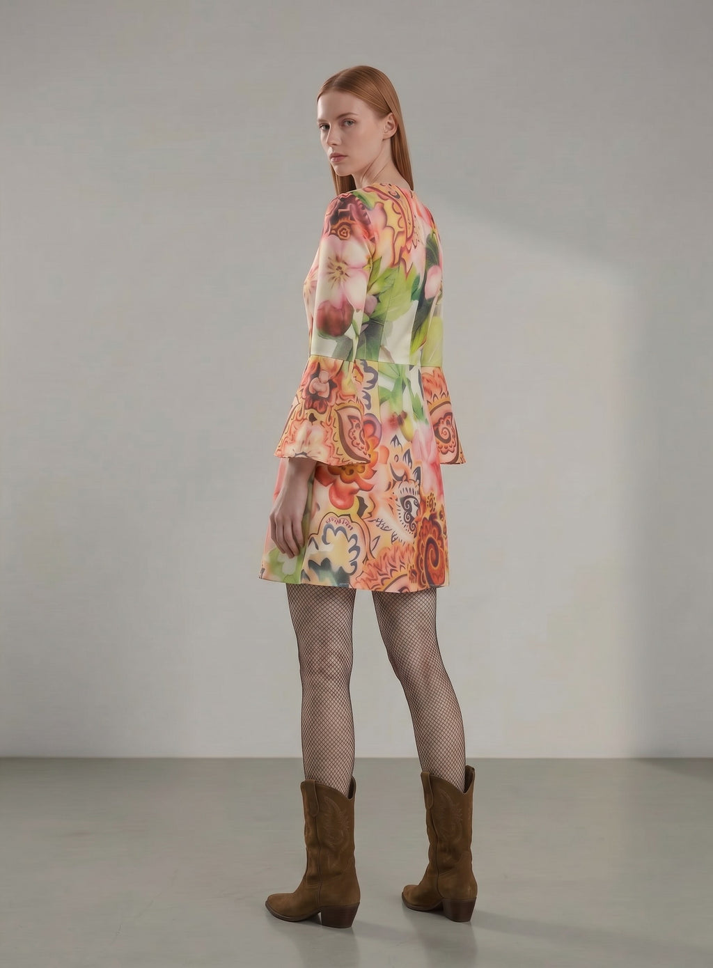 Day One Concept Store A-line mini dress with floral and paisley print, bell sleeves, and round neckline. Model wears fishnet tights and brown boots.